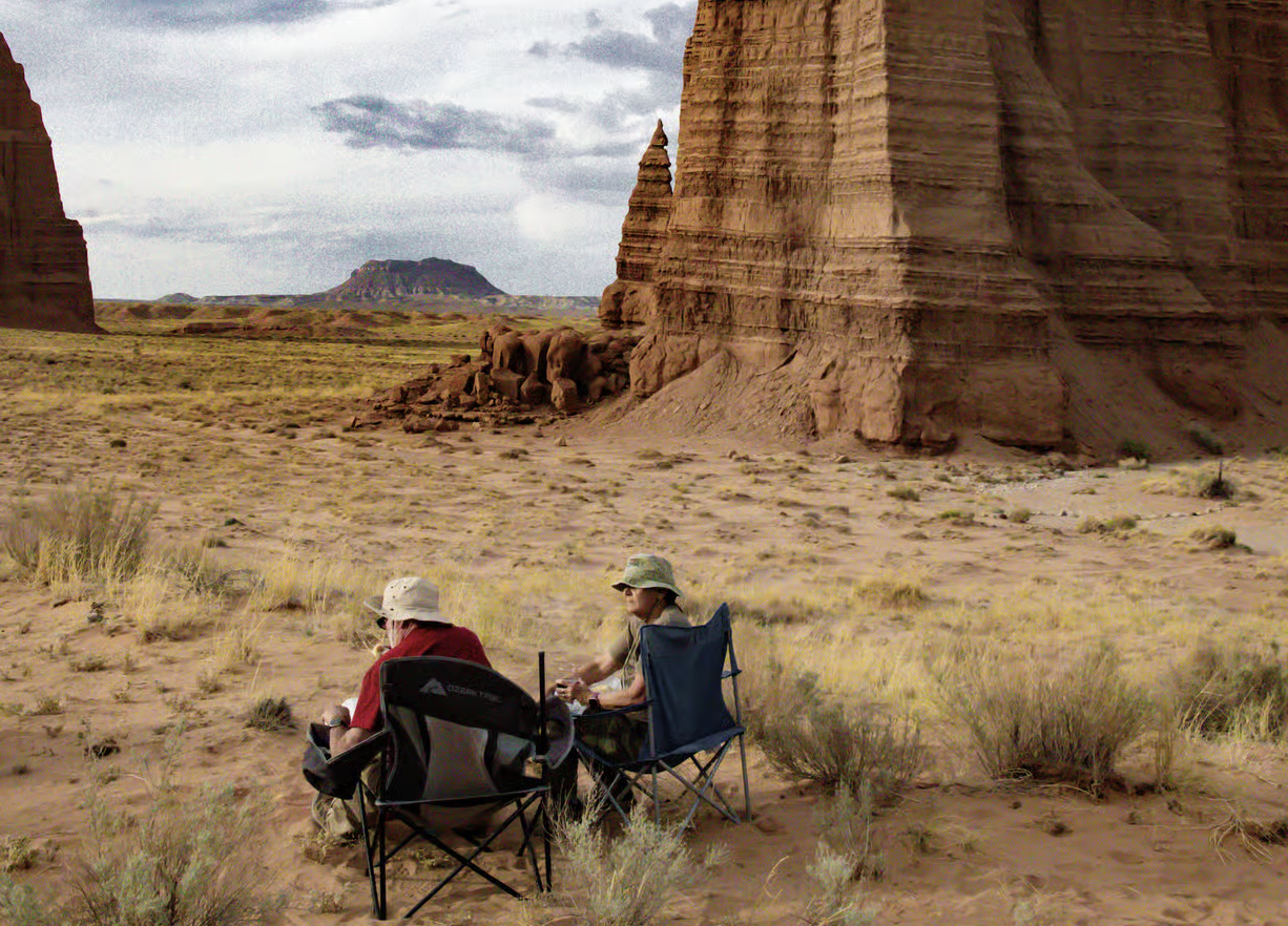 Two people in camp chairs viewing distant red rock monoliths across a grassy desert valley