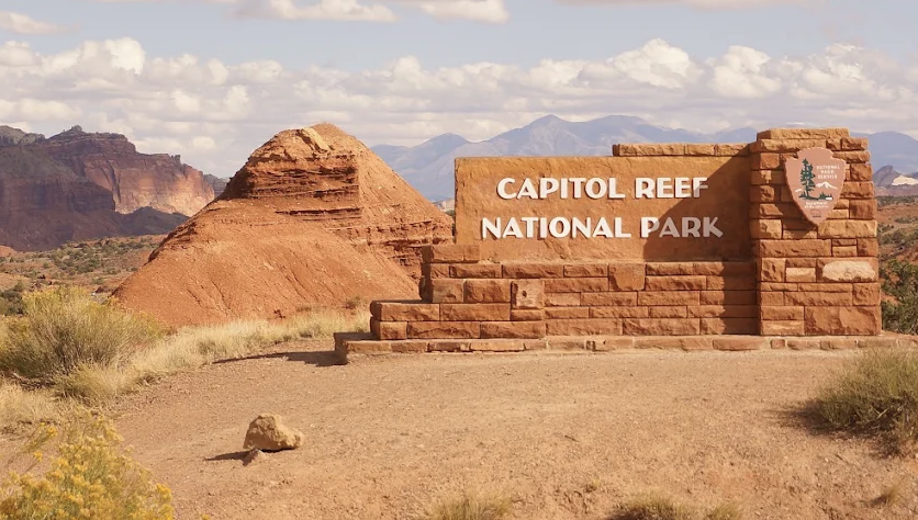 Capitol Reef National Park entrance sign with a rounded red butte and distant mountains under a daytime sky