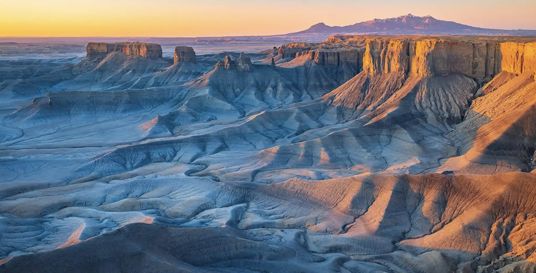 Factory Butte glowing orange at sunset above gray badlands with mountains behind, outside Capitol Reef National Park