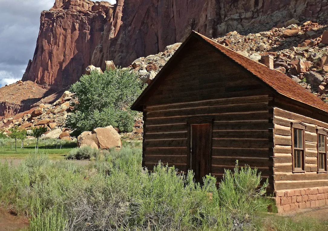 The one room log Fruita Schoolhouse with red cliffs behind in Capitol Reef National Park