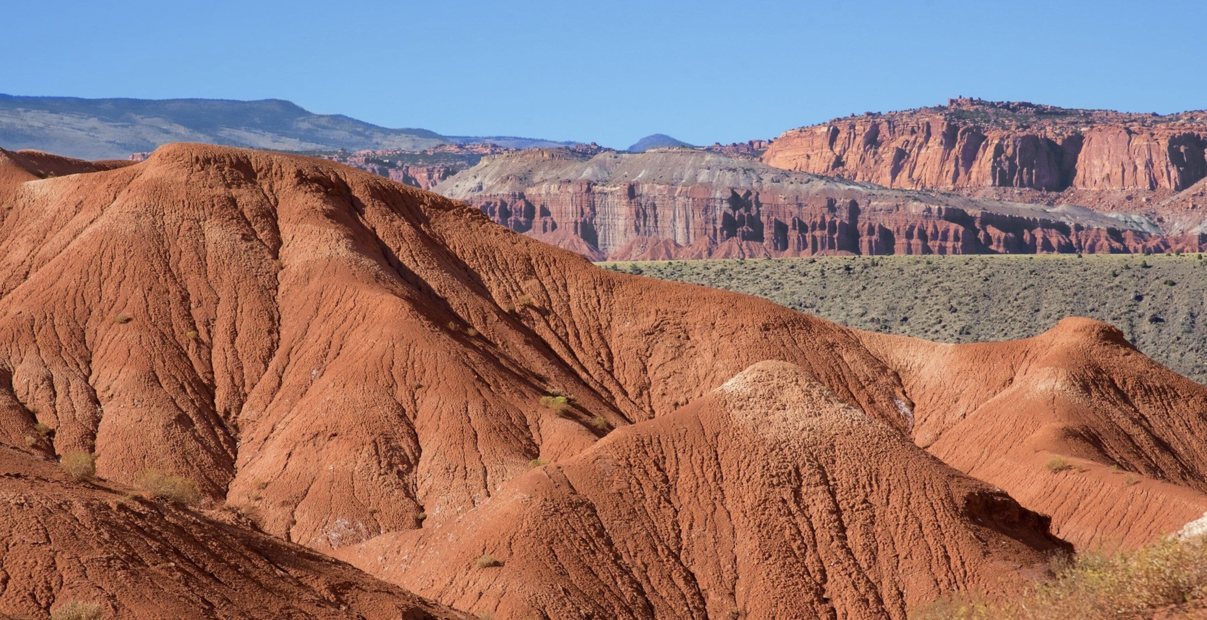 Smooth red badlands mounds with the reef's banded cliffs and distant mountains behind in Capitol Reef National Park
