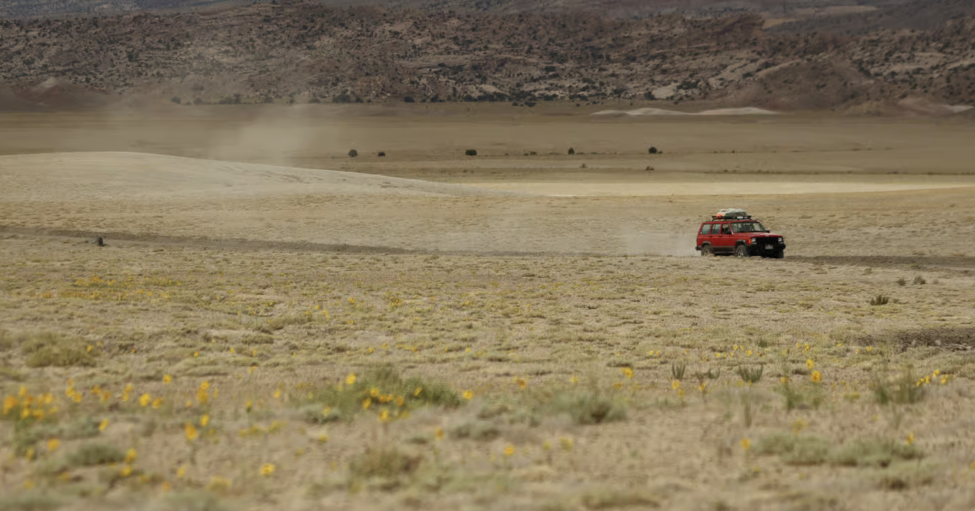 A red SUV kicking up dust while crossing a flat desert playa with wildflowers in the foreground