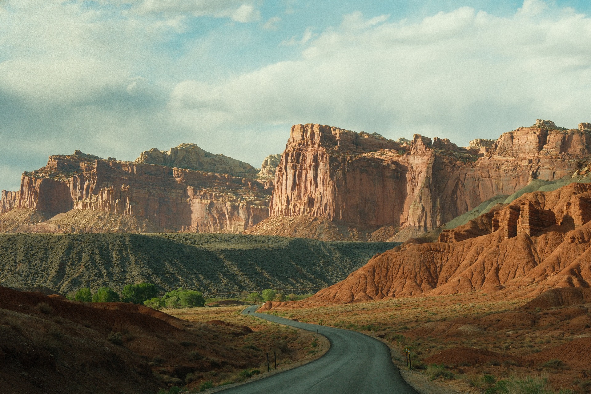 Two lane road winding through red and tan sandstone cliffs under a partly cloudy sky