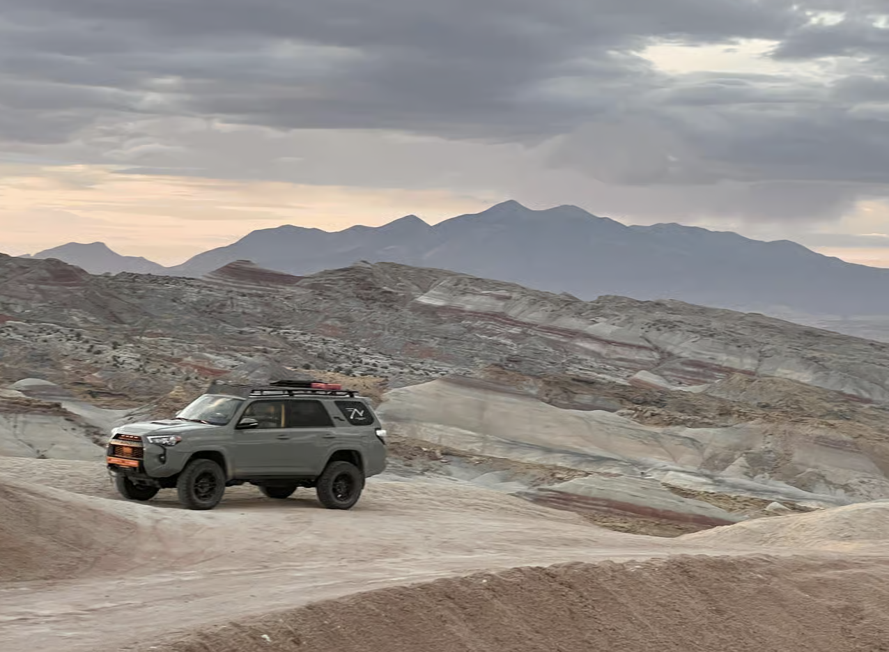 A gray SUV parked on a dirt road above banded gray and red badlands with mountains behind, near Capitol Reef