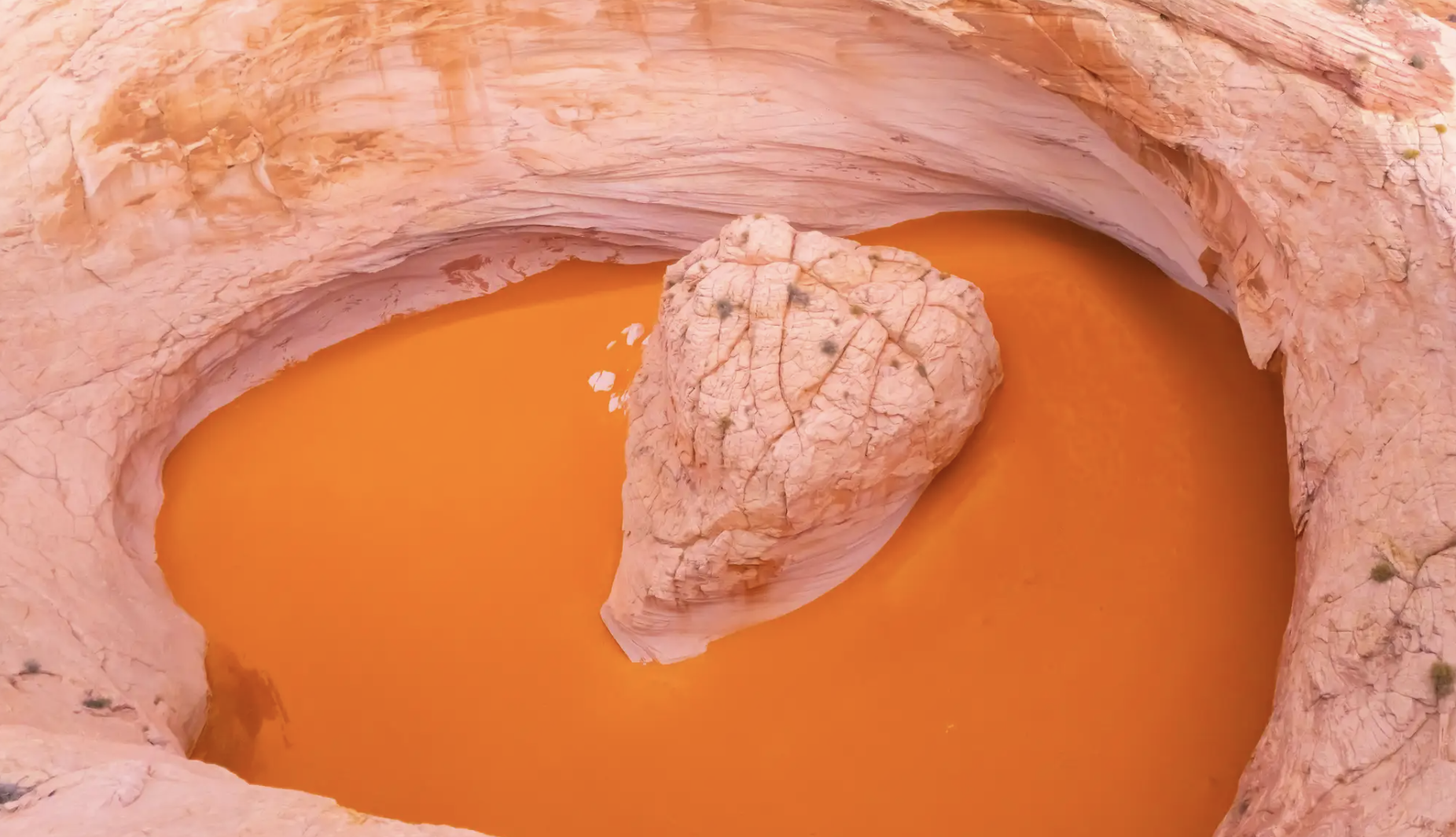 A sandstone waterpocket filled with vivid orange water surrounding a pale boulder